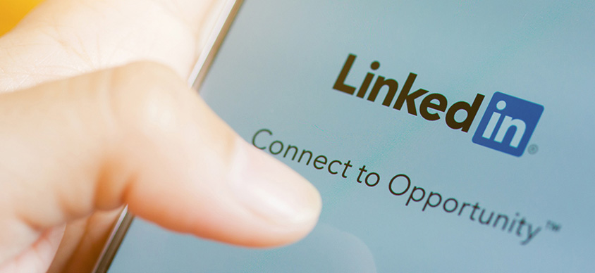 Un telefono cellulare con schermata che mostra il logo della piatatforma linkedIn dove il dito di un utente sta cliccando per accedere alla piattaforma di riferimento al fine di rappresentare il servizio di connessione e di opportunità lavorative che offre la piattaforma stessa